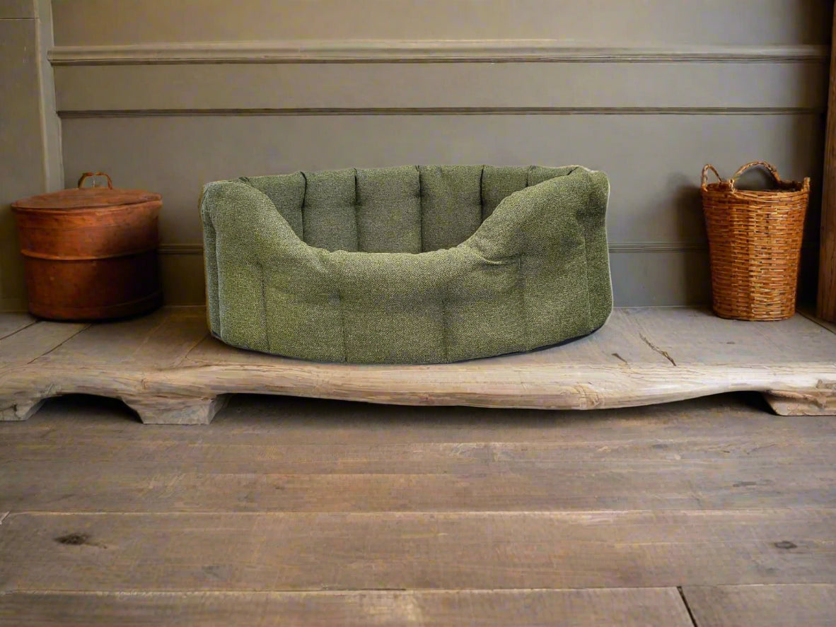 Green tweed oval dog bed with tufted cushion on wooden platform, flanked by wicker baskets in rustic setting