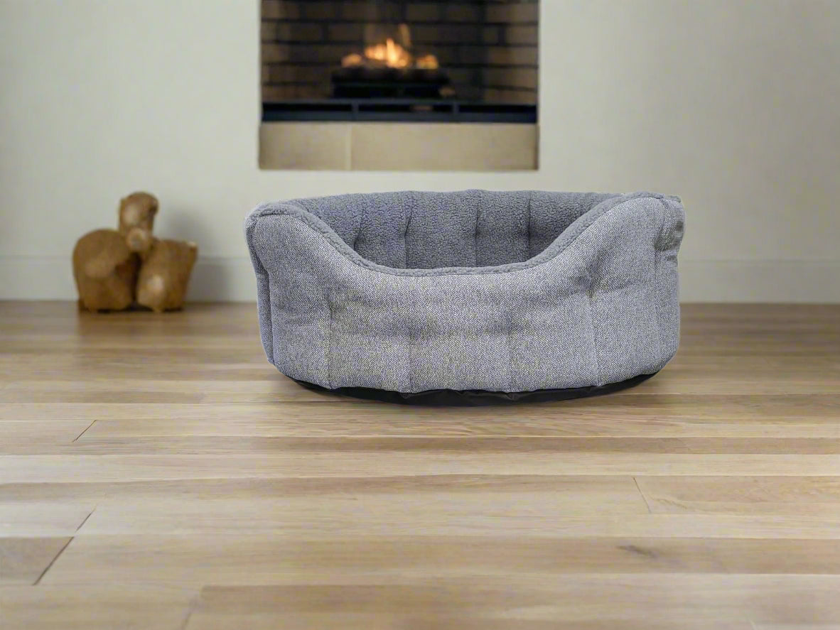 Grey tweed fleece lined dog bed with raised bolster edges on wooden floor near fireplace, cozy pet bedding