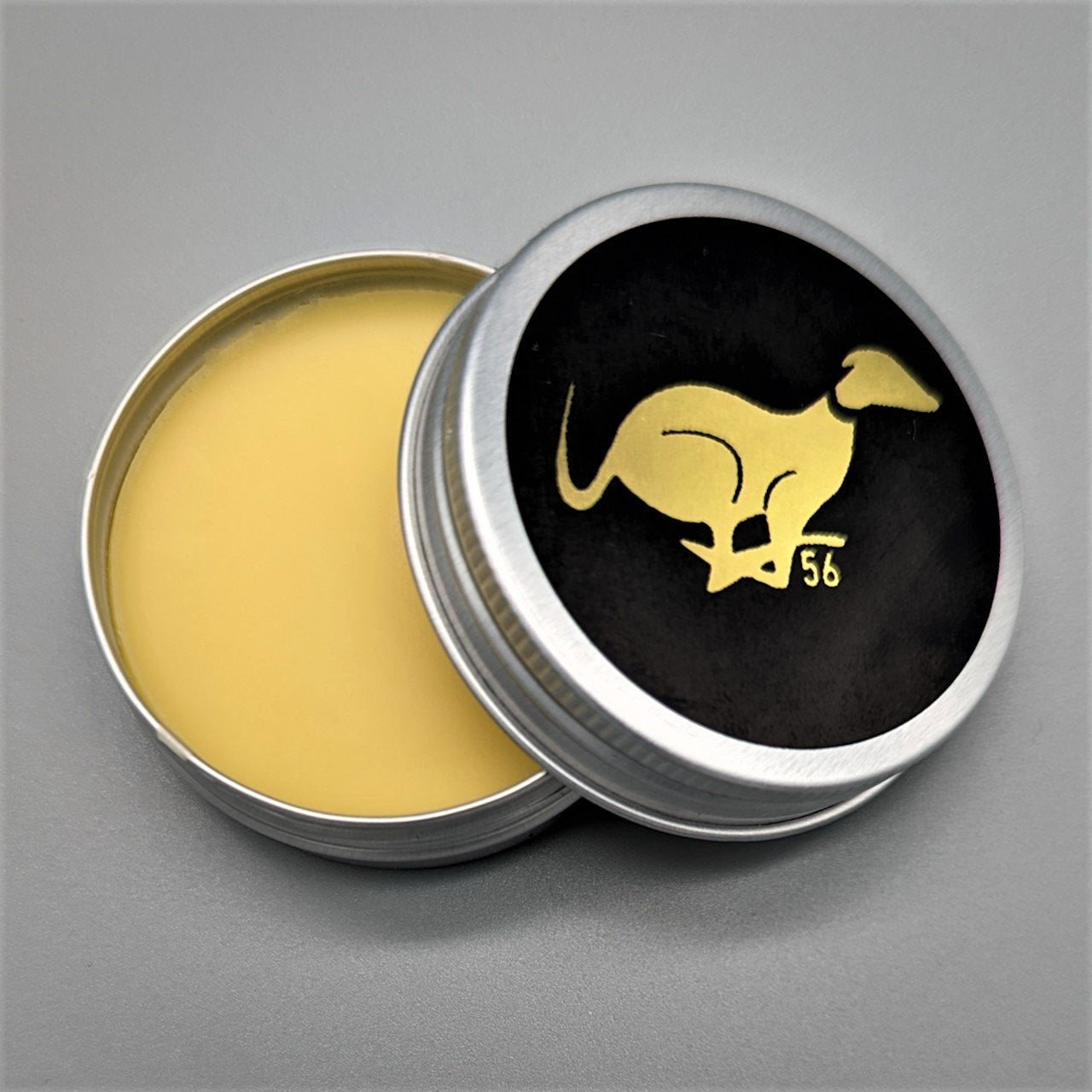 Open tin of premium all-natural leather balm with golden wax formula and elegant black lid featuring gold logo
