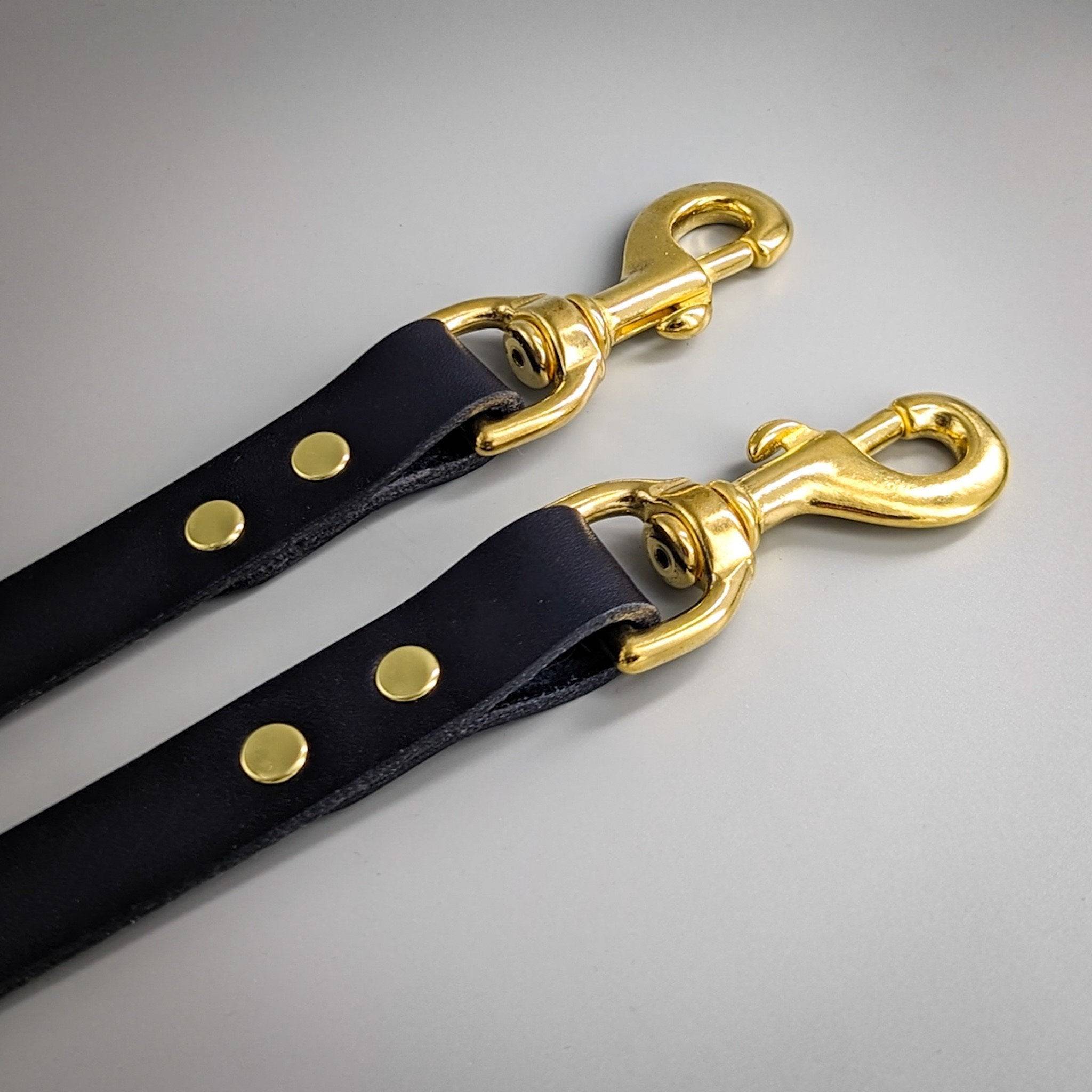 Two black leather dog training leads with polished gold metal hardware and brass rivets for adjustment
